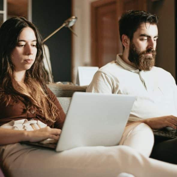 A woman with long dark hair and a bearded man sit together using separate laptops.