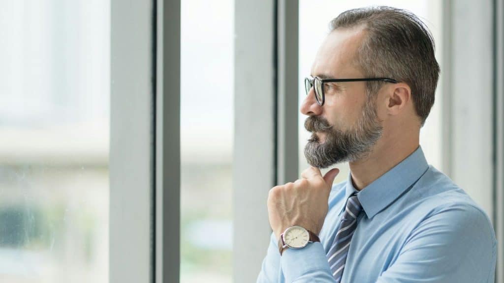 A man with a beard and glasses looks out a window with his hand on chin.