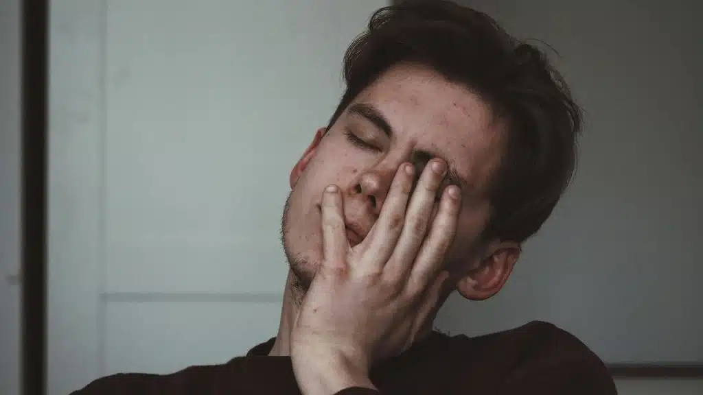 A person with eyes closed, resting their face in one hand against a plain background.