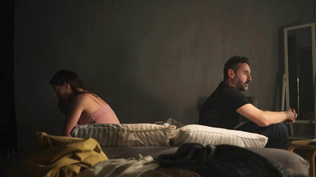 A couple sitting back to back on a bed in a dimly lit room.