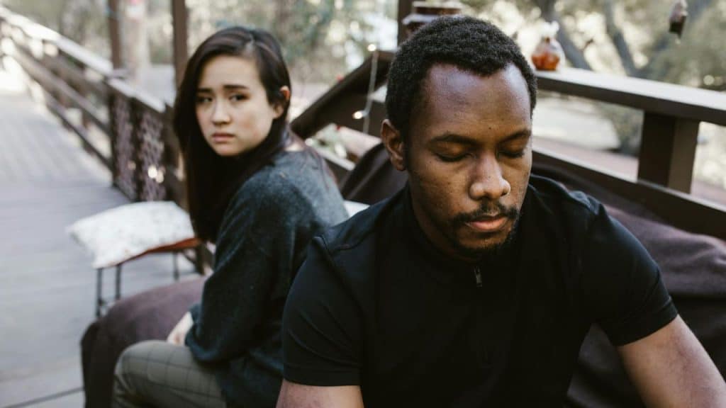 A man with closed eyes sits in the foreground while a woman looks on behind him.