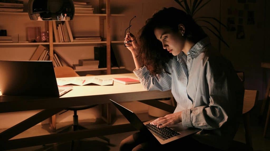 A woman working on a laptop at a desk, holding glasses in a dimly lit room.