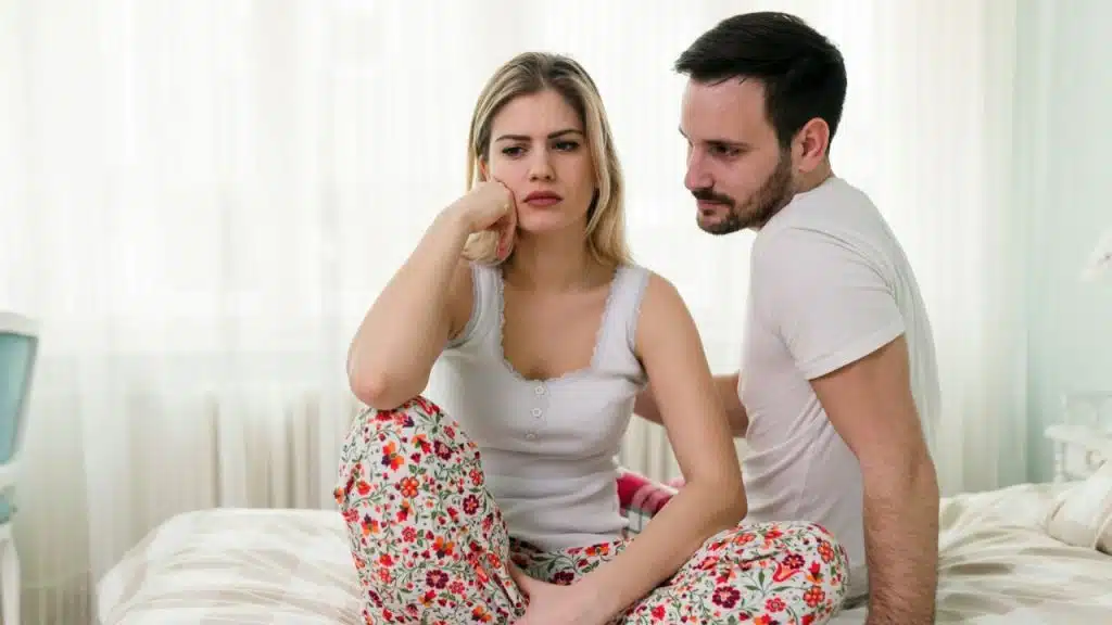 A man sits on a bed looking at a woman who is staring straight ahead.