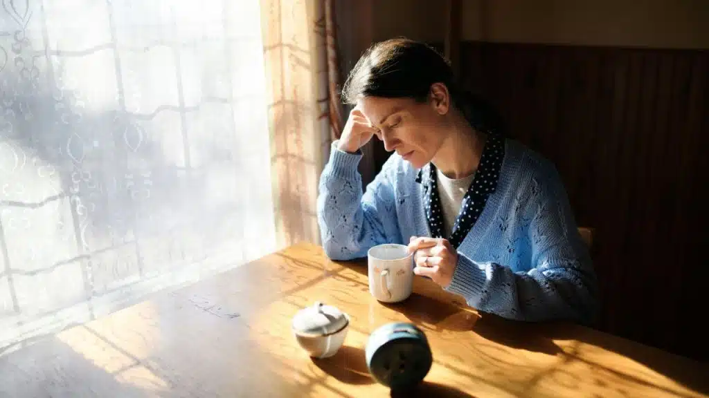 A woman in a blue cardigan sits at a wooden table looking down at a mug.