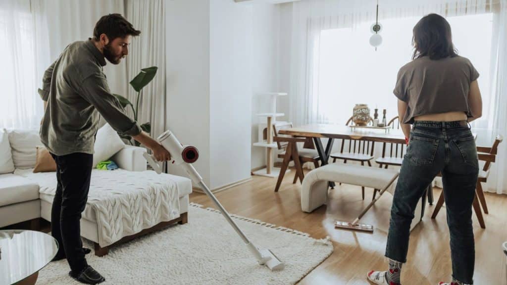A man vacuums a white rug while a woman mops a wooden floor in a living room.
