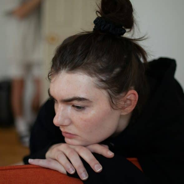 A woman resting her chin on her hands, that rest on the head of a couch which she sits on.
