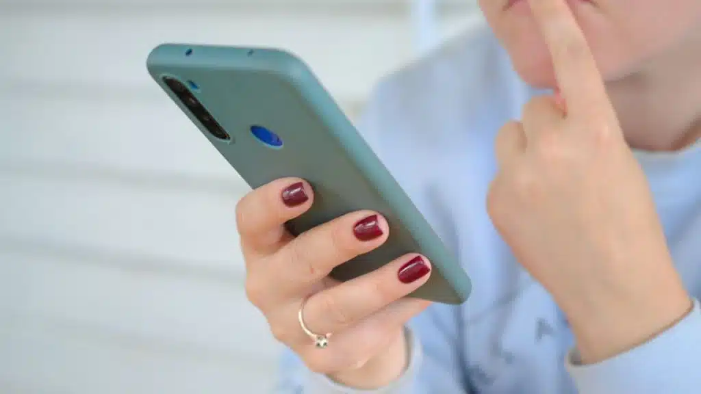 A person with dark red nail polish holds a green smartphone and touches their chin.