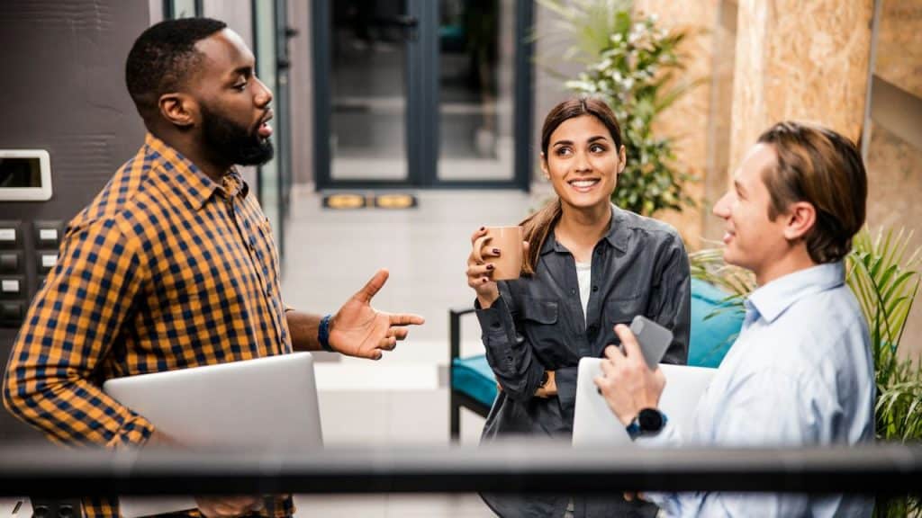 Three people stand in a modern office hallway holding coffee mugs and laptops while talking.