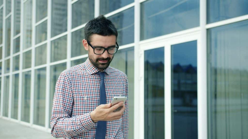A man with glasses and a tie looks at his phone in front of glass panels.