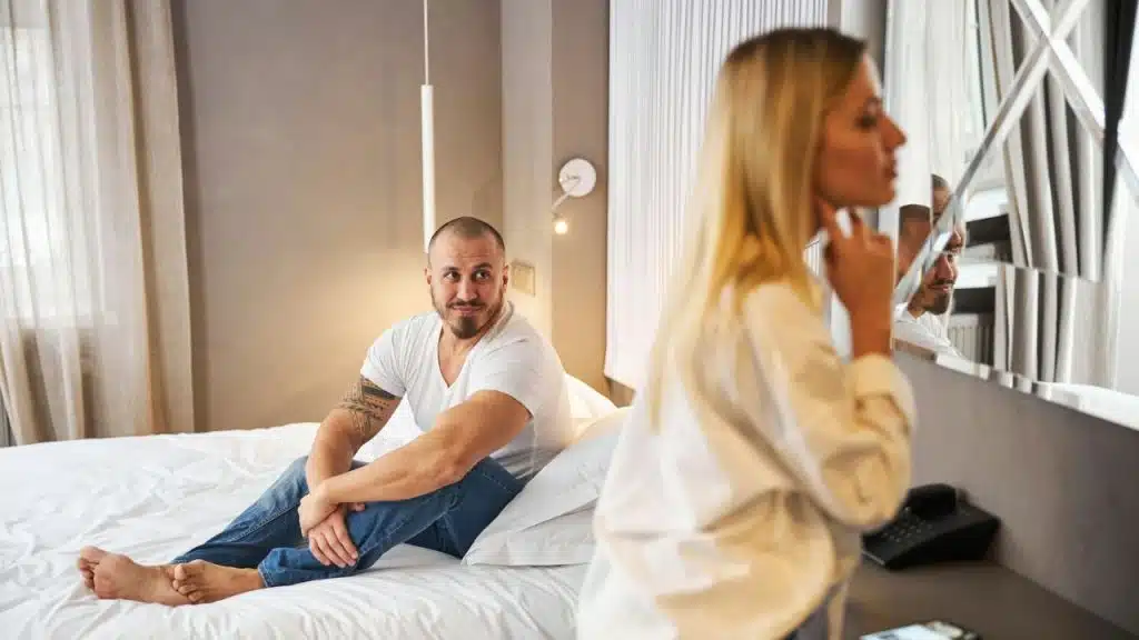 A man sits on a bed looking toward a woman who is looking in a mirror.