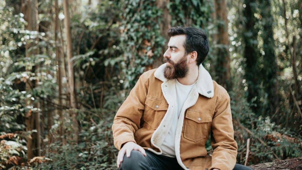 A bearded man in a sherpa-lined tan jacket sits in a forest looking to his right.