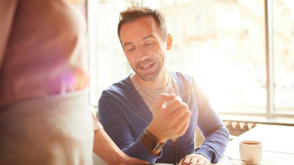 A man smiling as he chats with someone at a sunlit café table.
