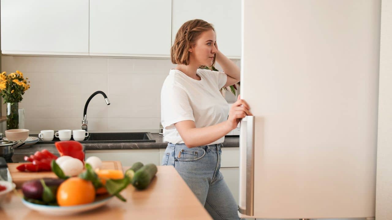 A woman stands in a kitchen looking into an open white refrigerator near fresh produce.