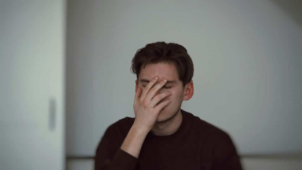 A young man covering his face with his hand, sitting indoors against a plain wall.