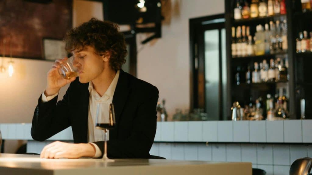 A man in a suit drinks from a glass while sitting alone at a bar counter.