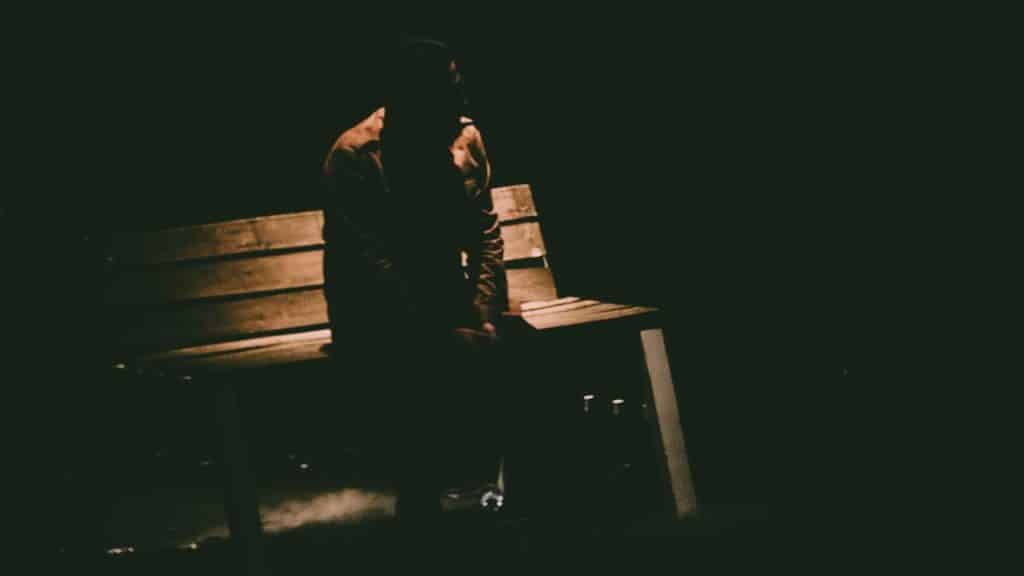 A lone figure sits on a wooden bench in a dark and shadowed outdoor setting.