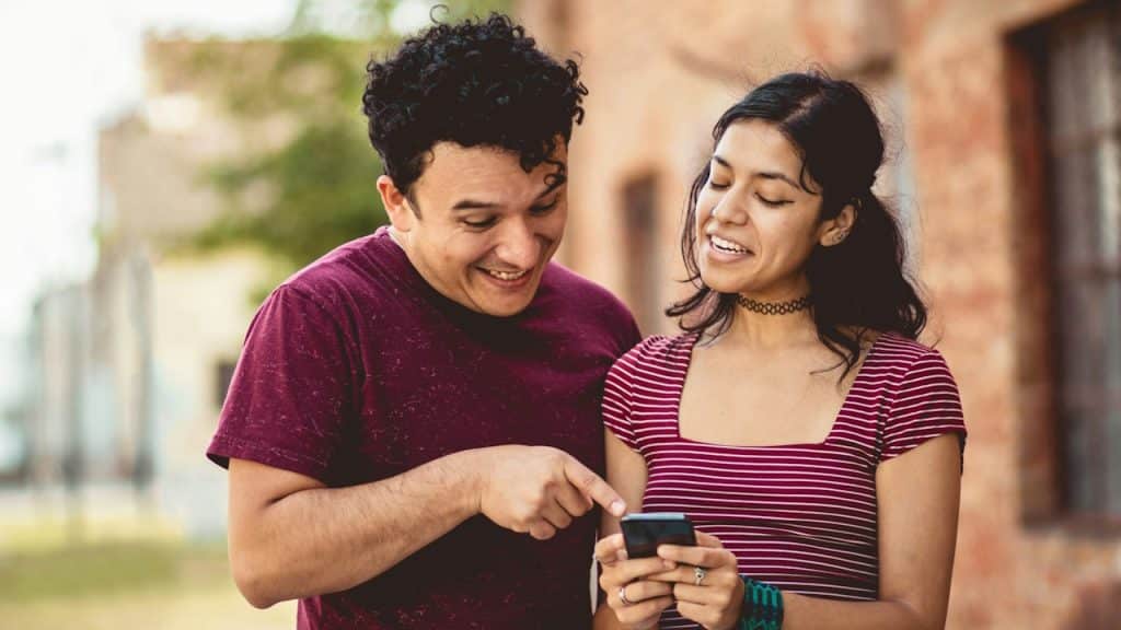 A young smiling couple using their phones while they are standing in a street.