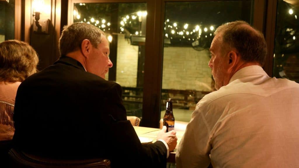 Two men in formal attire sit at a dimly lit restaurant table, talking near a window.