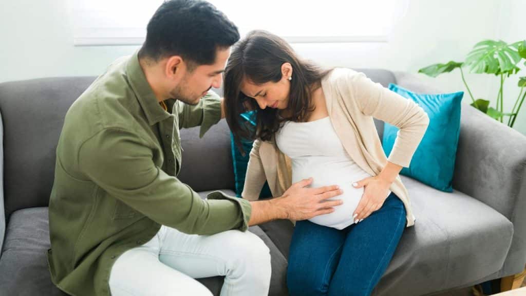 A couple sitting on a couch with the man gently touching a pregnant belly.