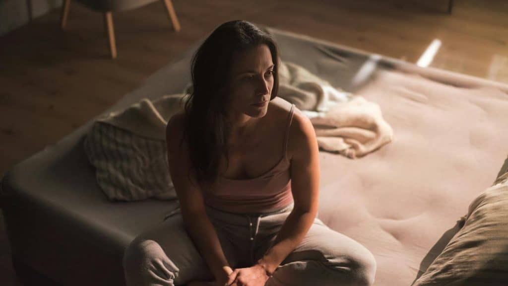 A woman sitting on a bed in soft light, looking off thoughtfully.