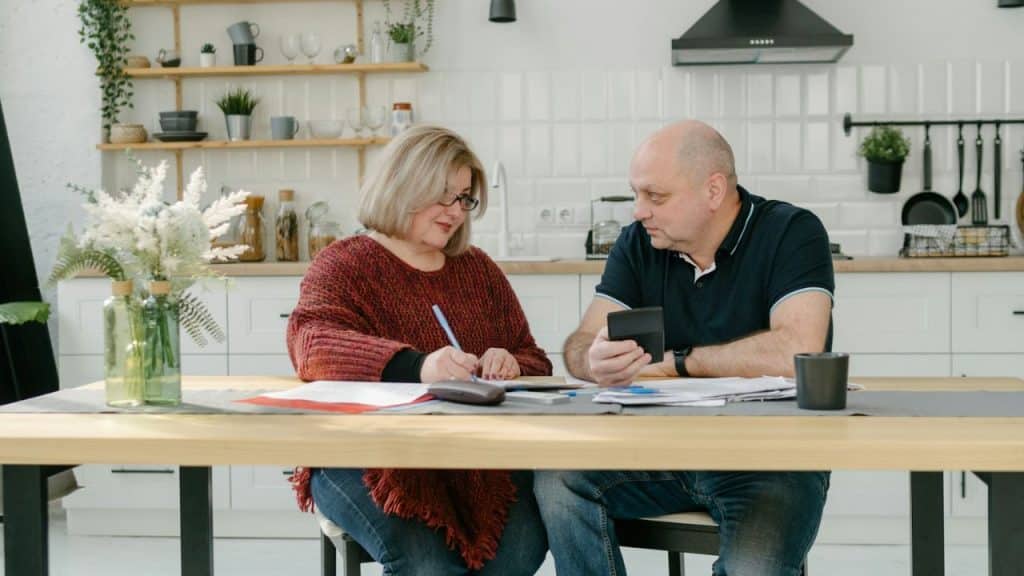 A man and woman sit at a table in a kitchen reviewing papers and a calculator.