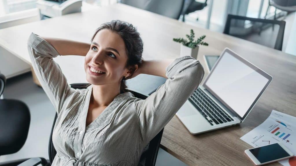 A smiling woman leaning back in an office chair beside a laptop and documents.