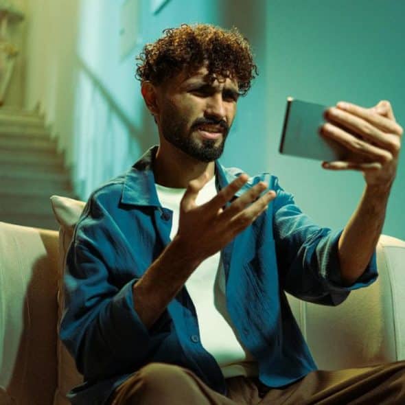 A man with curly hair sits on a sofa, gesturing while holding a smartphone upwards.