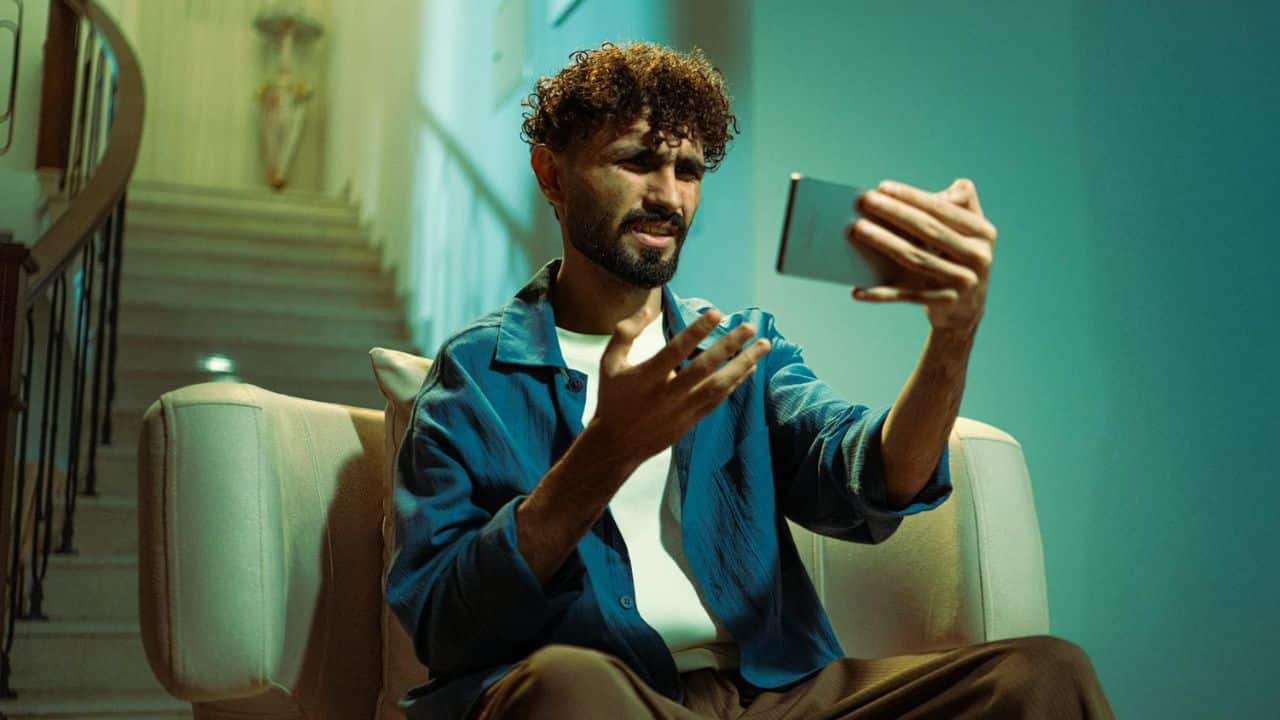 A man with curly hair sits on a sofa, gesturing while holding a smartphone upwards.
