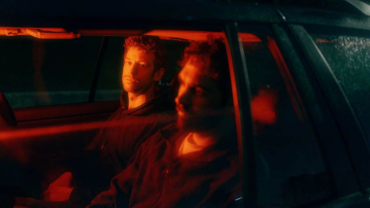Two men sit in a dark car illuminated by a red light from the side.