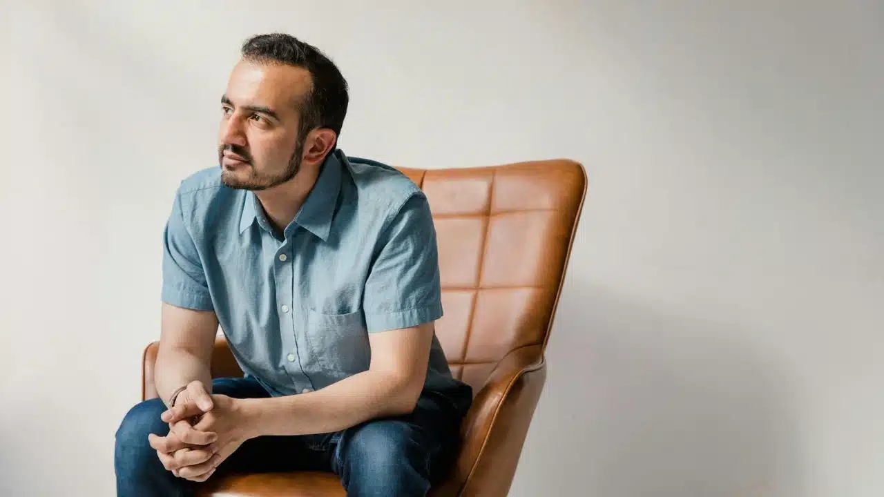 A man in a blue shirt sits in a brown leather chair, looking to the side.