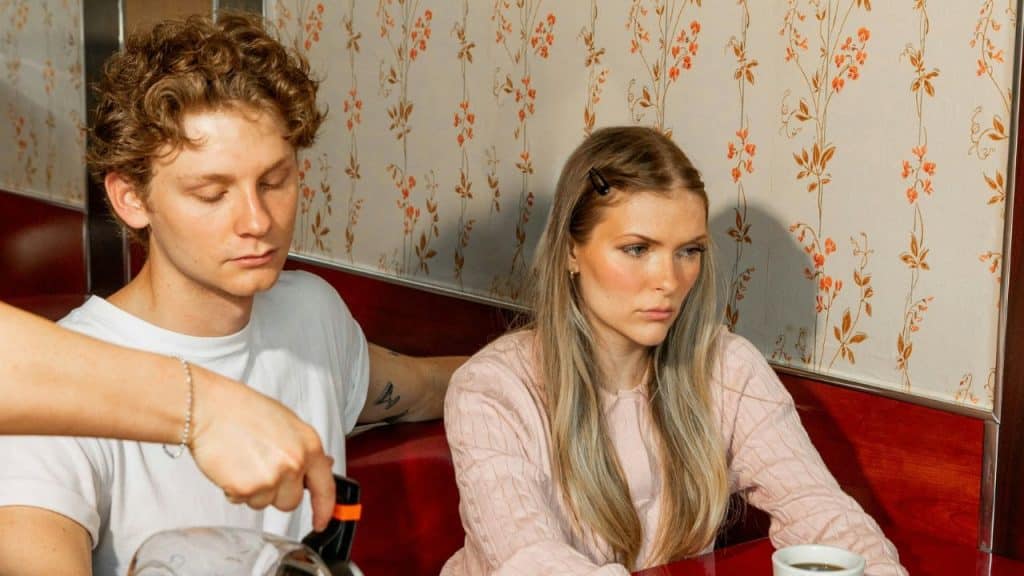 A young man and woman sit in a booth while someone pours coffee into a cup.