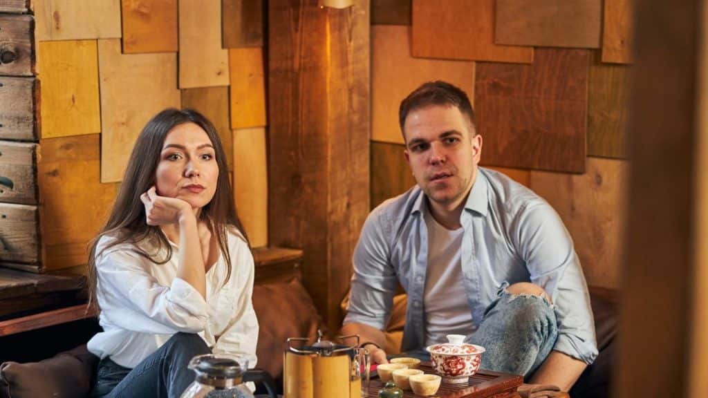 A man and woman sit together in a room with wood-paneled walls and a tea set.