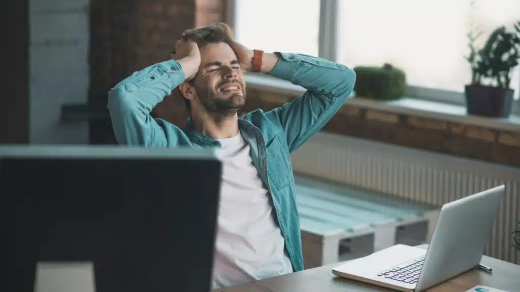 A man in a denim shirt sits at a desk with his hands on his head.