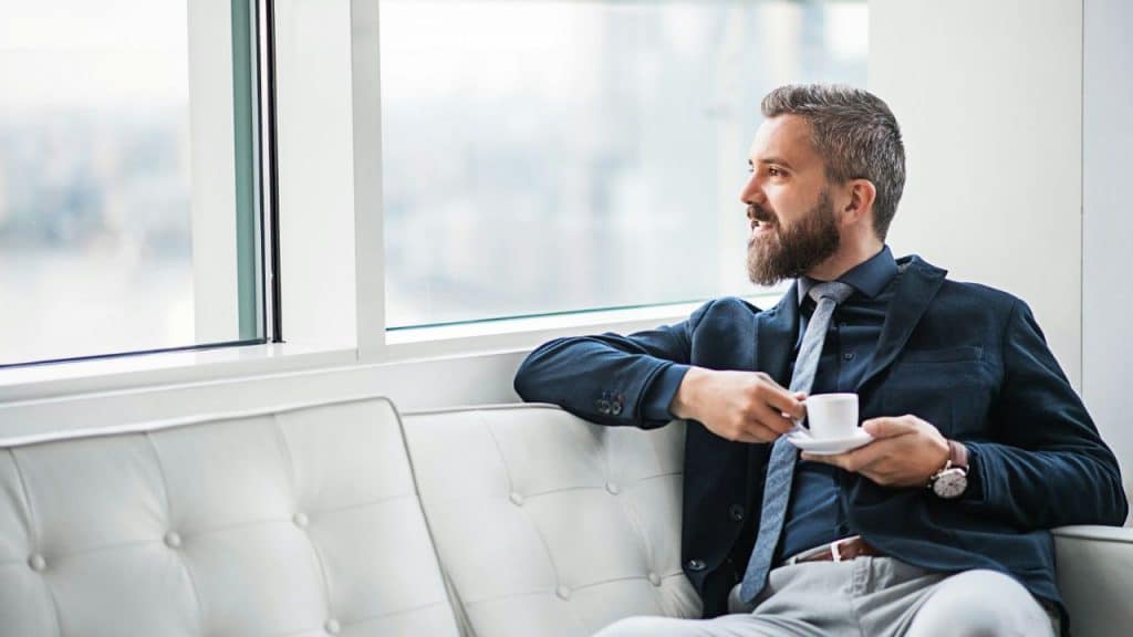A bearded man in a suit sits on a white sofa, smiling out a window.
