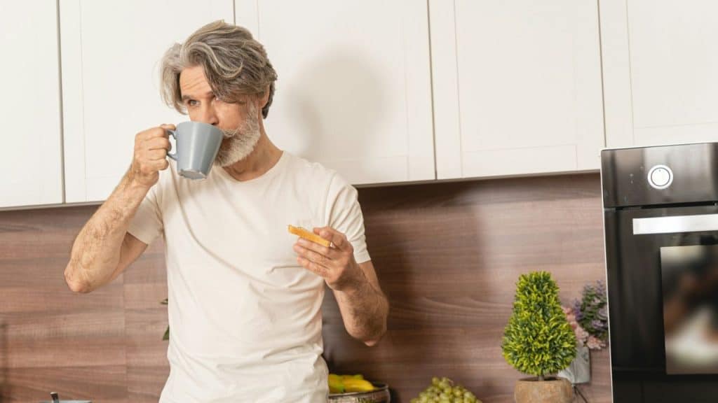 A man with silver hair and a beard drinks from a gray mug in a kitchen.