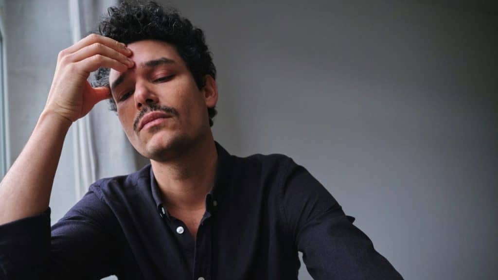 A man with curly hair and a mustache sits with his hand on his forehead.