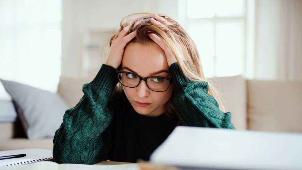 A woman wearing glasses and a green sweater holds her head in her hands while looking down.