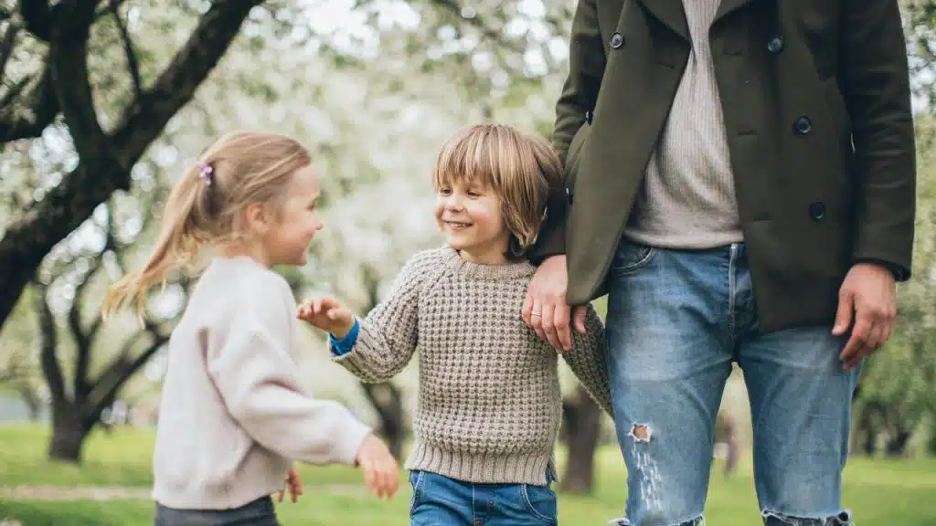 A small child holding an adult’s hand while smiling at another child in a park.Even a throwaway line about how Lena’s husband always helps with the dishes, or Chris planned a whole weekend for his wife, can leave a mark.