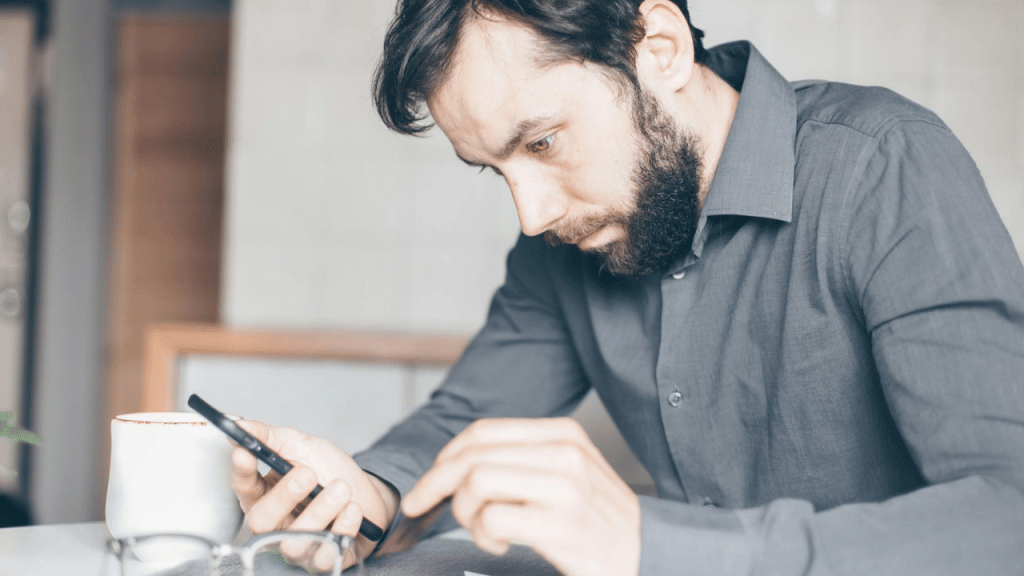 A bearded man in a gray collared shirt looks down at a smartphone in his hand.