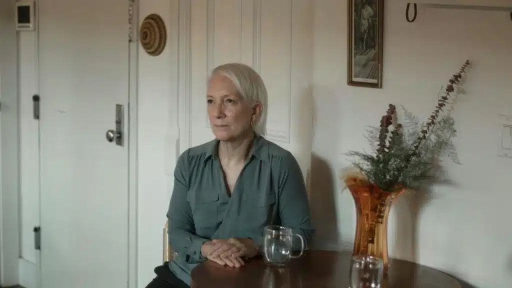 An older woman sitting at a table with a glass of water and a vase of flowers.
