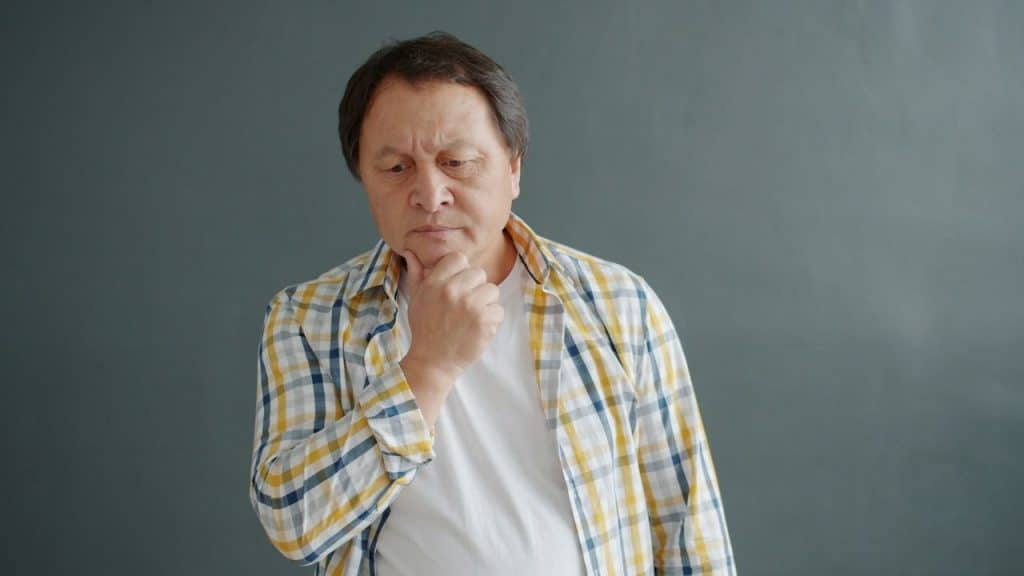 A man in a plaid shirt stands with his hand on his chin, looking downward pensively.