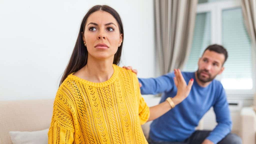 A woman in a yellow sweater looks away while a man reaches toward her shoulder.