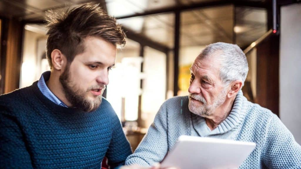 An older man and a younger man sit together looking at a white digital tablet.