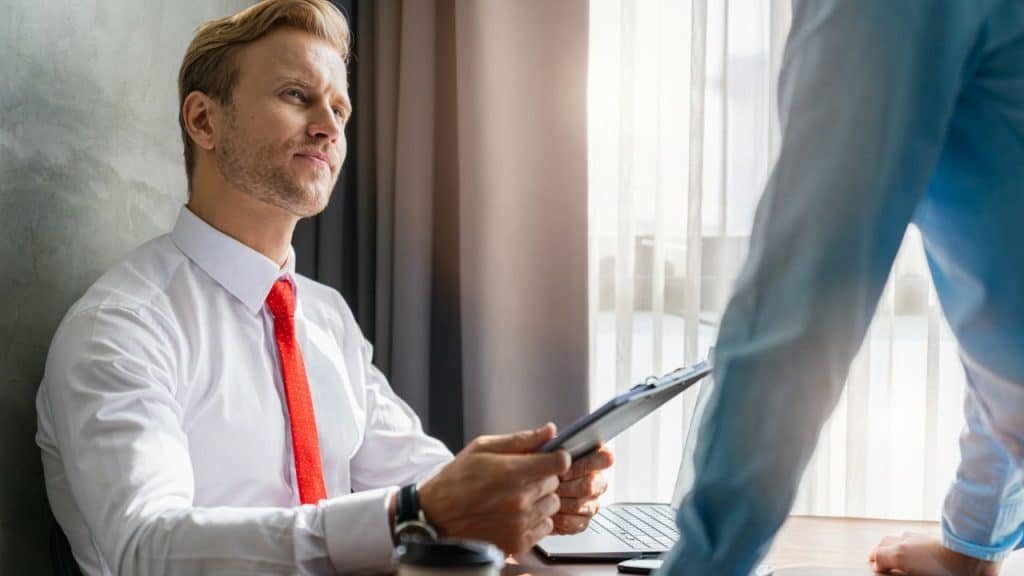 A blonde man in a white shirt and red tie looks up at another person.