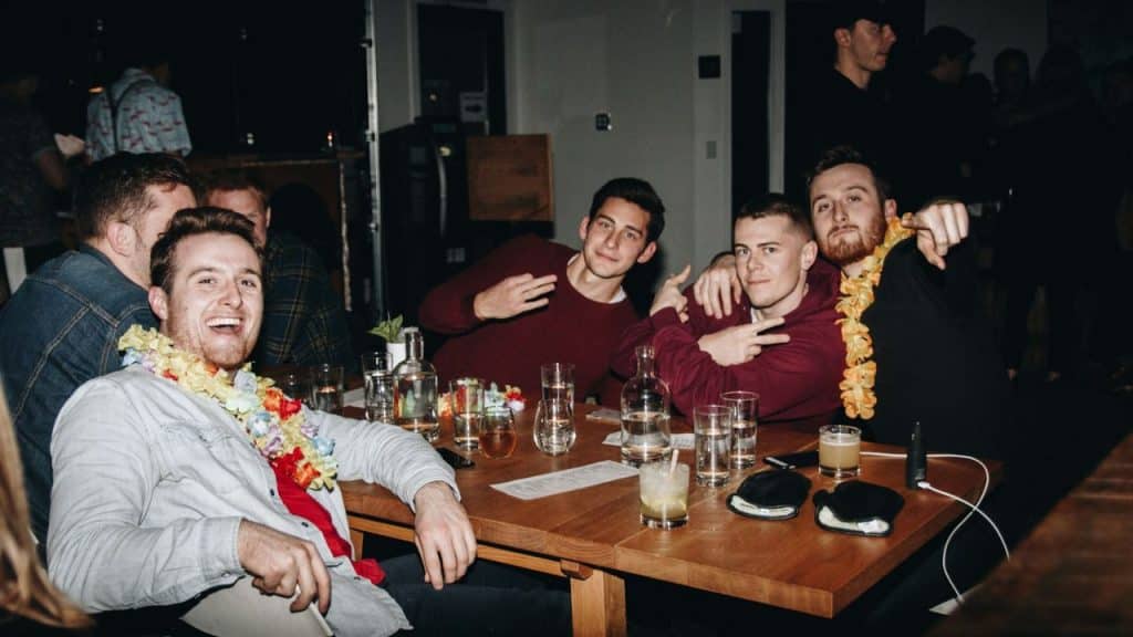 Six men sit at a wooden table in a dimly lit room with drinks.