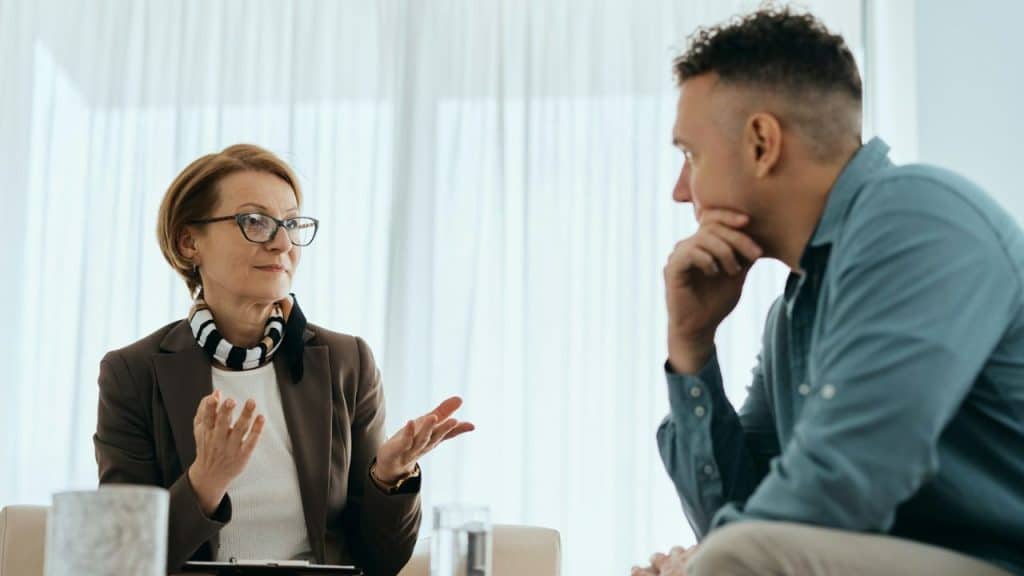A woman in a blazer gestures while speaking to a man who is listening intently.