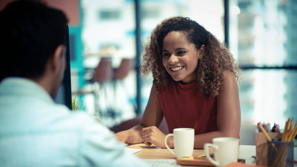 A smiling woman with curly hair sits at a table and talks to a man.