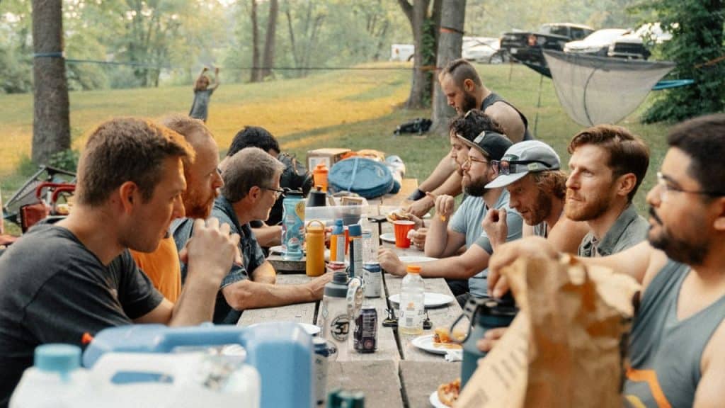 A group of men sit at a long wooden picnic table outdoors, eating and talking together.