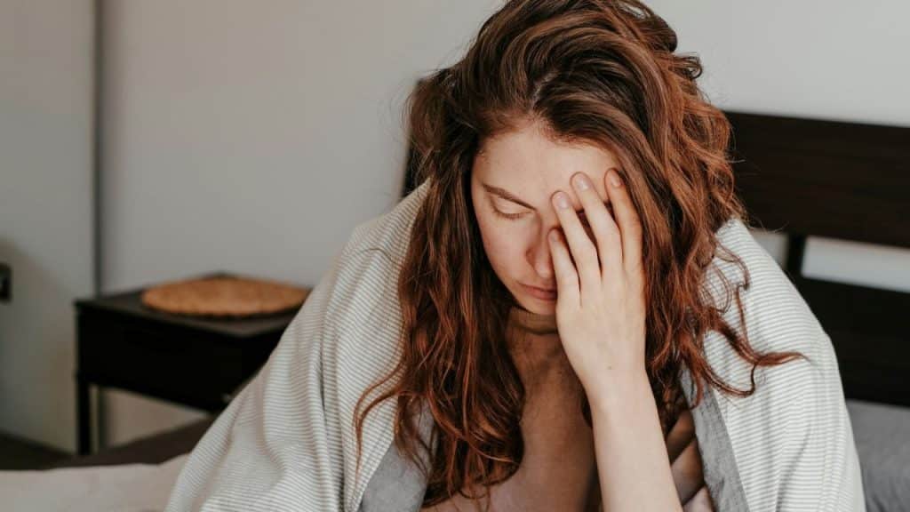 A woman with long, wavy brown hair is sitting up in bed, covering her face with one hand in distress.