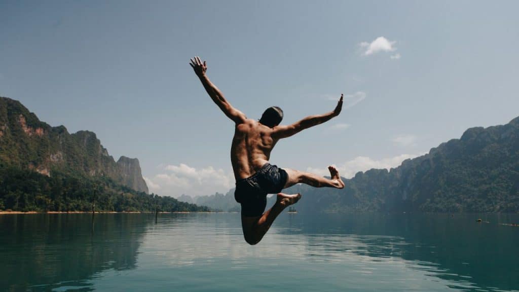 A shirtless person in swim trunks jumps into a body of water with mountains behind.